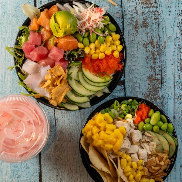 Two colorful Poki Bowls with various toppings and a pink drink on a distressed blue wooden surface.