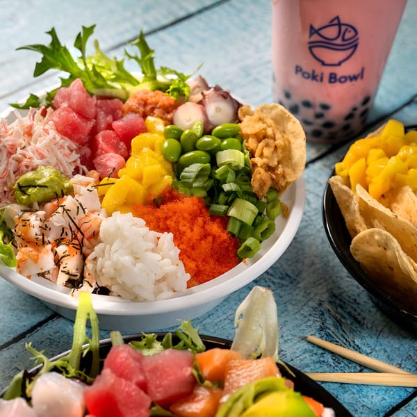Two colorful Poki Bowl poke bowls with a pink boba tea and chips on a blue wooden table.