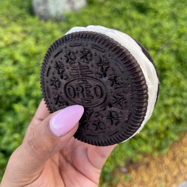 A hand with pink nails holds an Oreo ice cream sandwich against a green background.